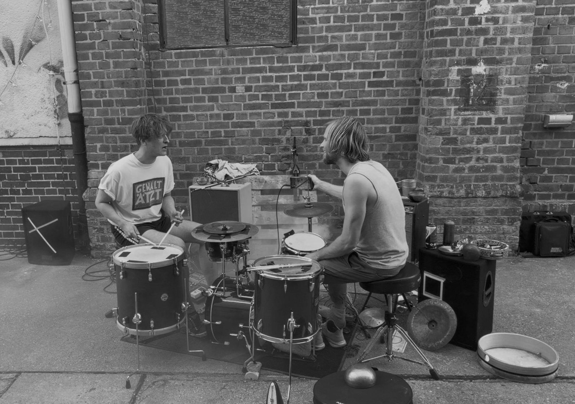 Martin Rother and Tom Schäfer facing each other at the drums, outdoors in front of a brick wall